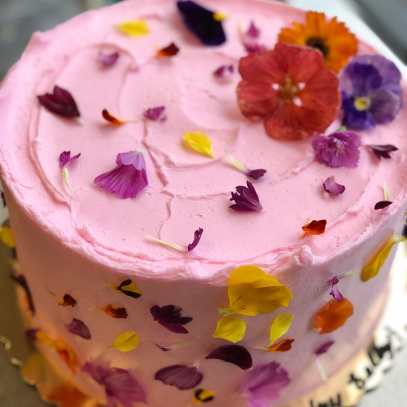 A round pink frosted cake adorned with colorful petals on top and around its sides.