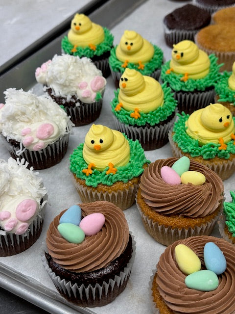 A tray of colorful Easter cupcakes, decorated as chicks, sheep, and nests with eggs.